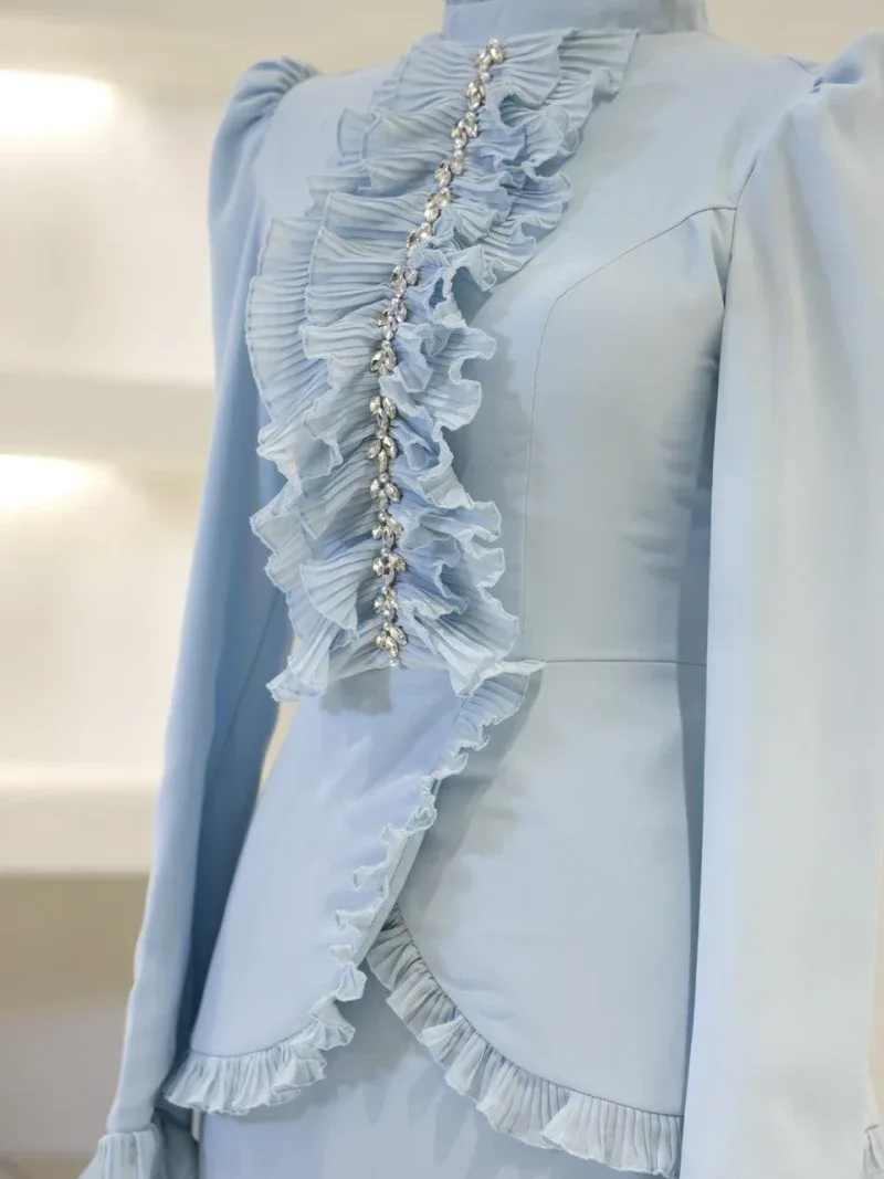Soft blue fabric blouse featuring stylish ruffle details along the front, accentuated with sparkling jewelry embellishments for a chic and sophisticated look.