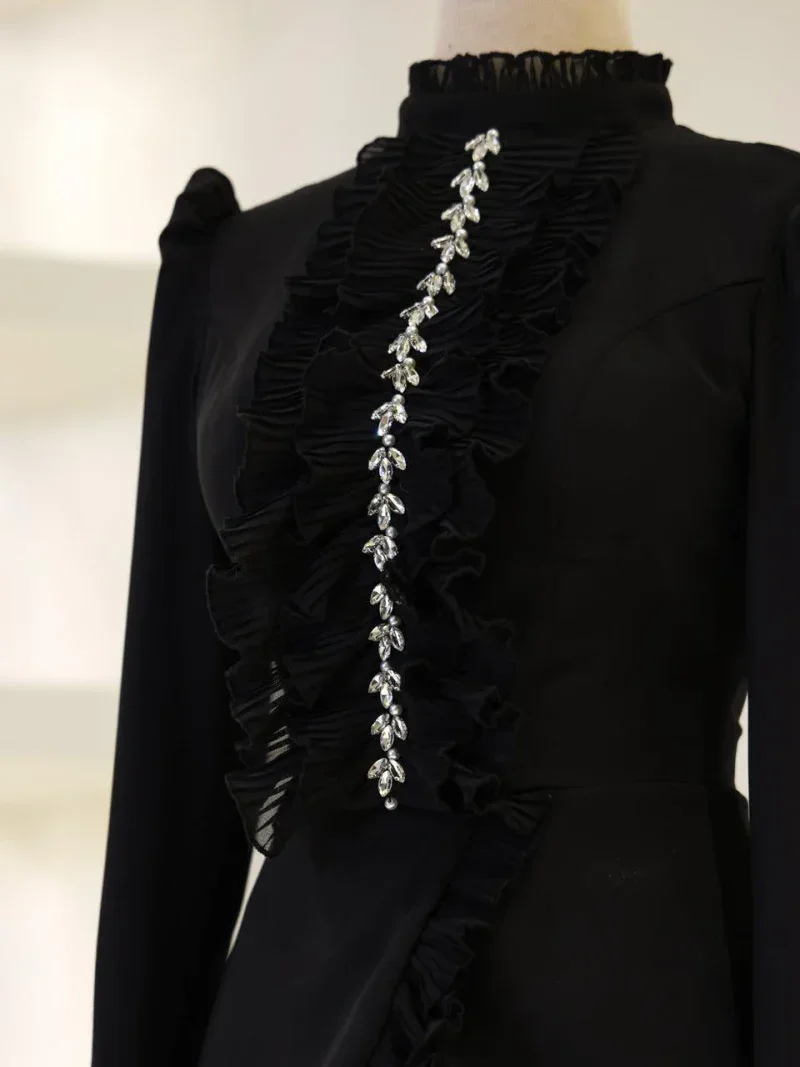 Beautiful black evening dress adorned with ruffled fabric and shiny crystal jewelry, ideal for parties, events, or special nights out. The high collar and detailed embellishments add a touch of sophistication and elegance to the attire.