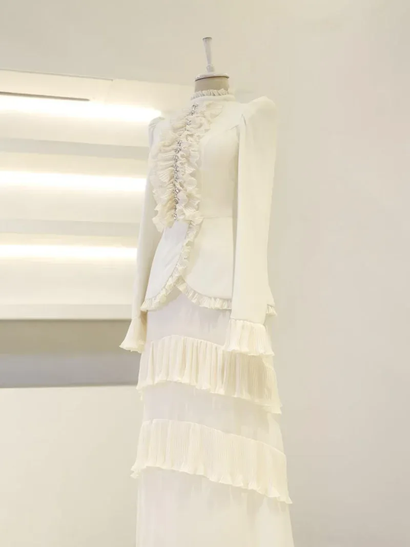 An elegant white dress featuring ruffles along the front and layered pleats at the bottom, displayed on a mannequin in a modern, minimalistic setting. Perfect for fashion enthusiasts and stylish occasions.