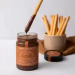 Rich chocolate hazelnut spread with a biscuit stick dipped in chocolate, on a white surface with a container of crispy breadsticks in the background.
