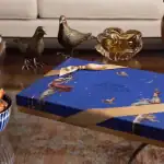 Elegant blue chocolate box with whimsical illustrations, wrapped with a satin ribbon, placed on a wooden table with decorative bird sculptures and a glass art piece in the background.