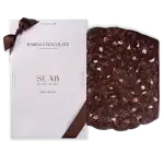 Rich dark chocolate with crushed toppings on a black background, paired with elegant white packaging featuring brown ribbon accents. Perfect for gourmet chocolate lovers.
